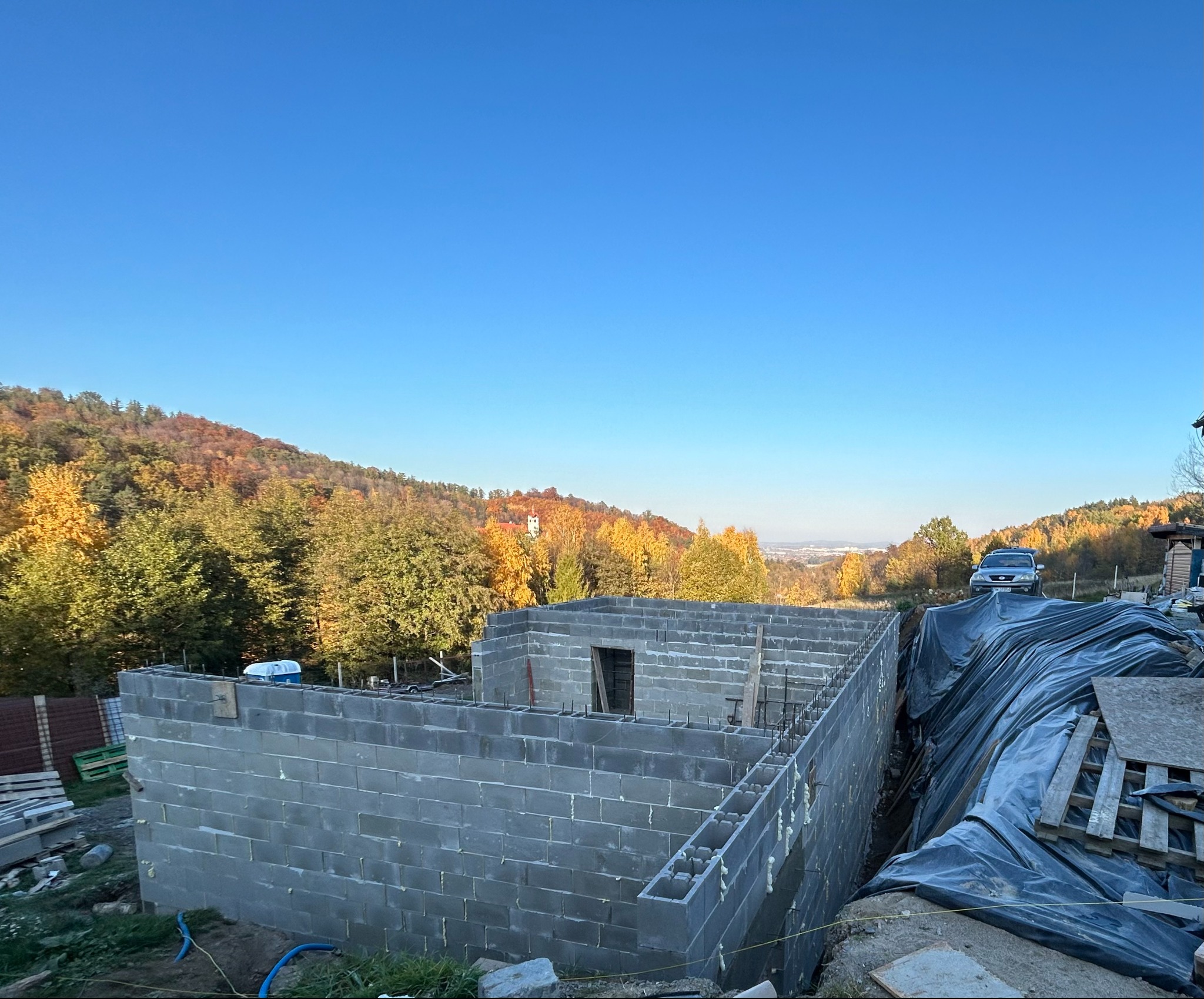 Wznoszone ściany budynku z bloczków betonowych na fundamencie, z widocznym zbrojeniem, w tle jesienny krajobraz zalesionych wzgórz i błękitne niebo.