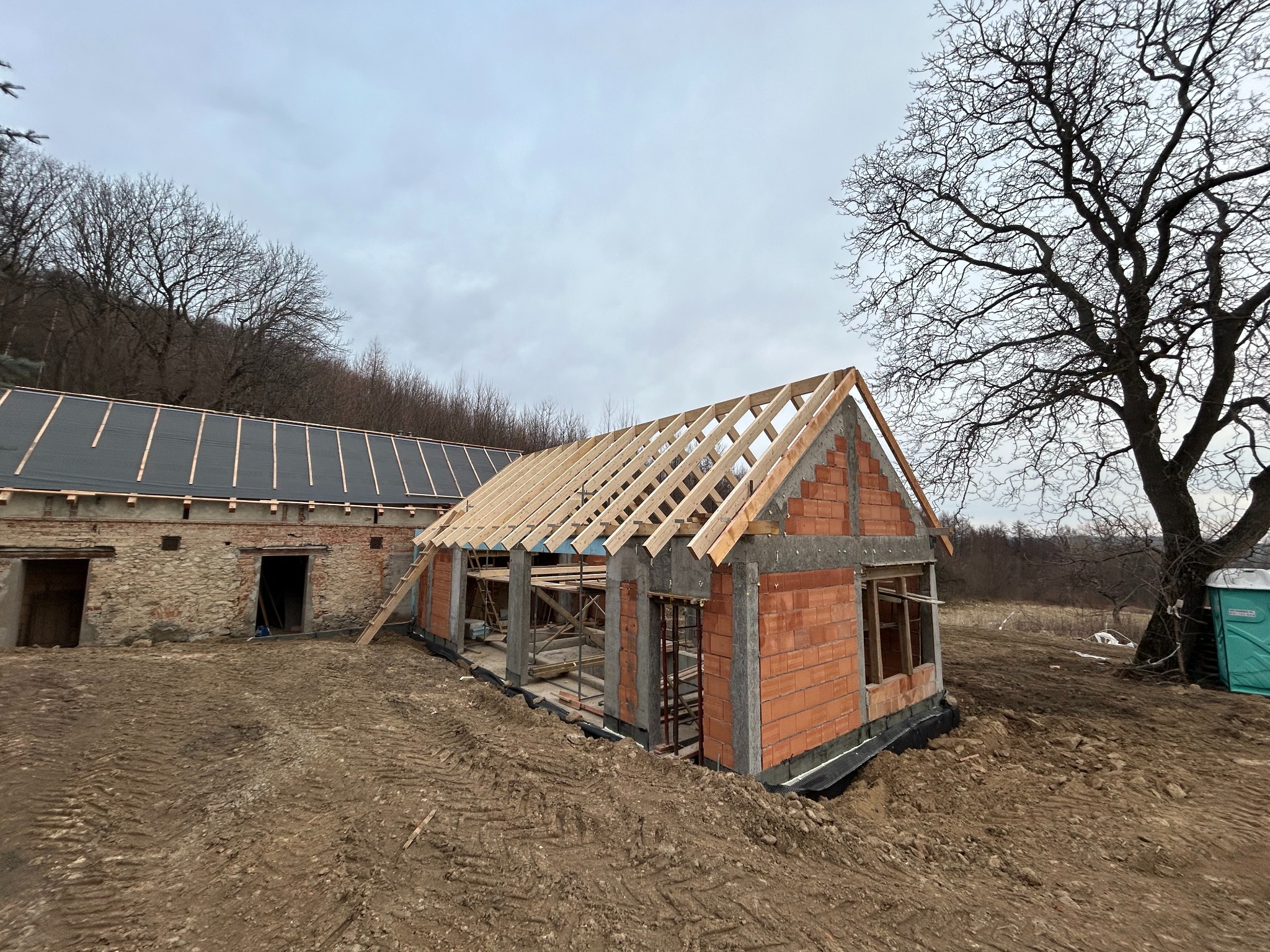 Budowa domu z czerwonej cegły, widoczne ściany z otworami okiennymi, drewniana konstrukcja dachu w trakcie montażu, obok budynek gospodarczy z ciemnym dachem, teren budowy z ziemią i śladami opon...