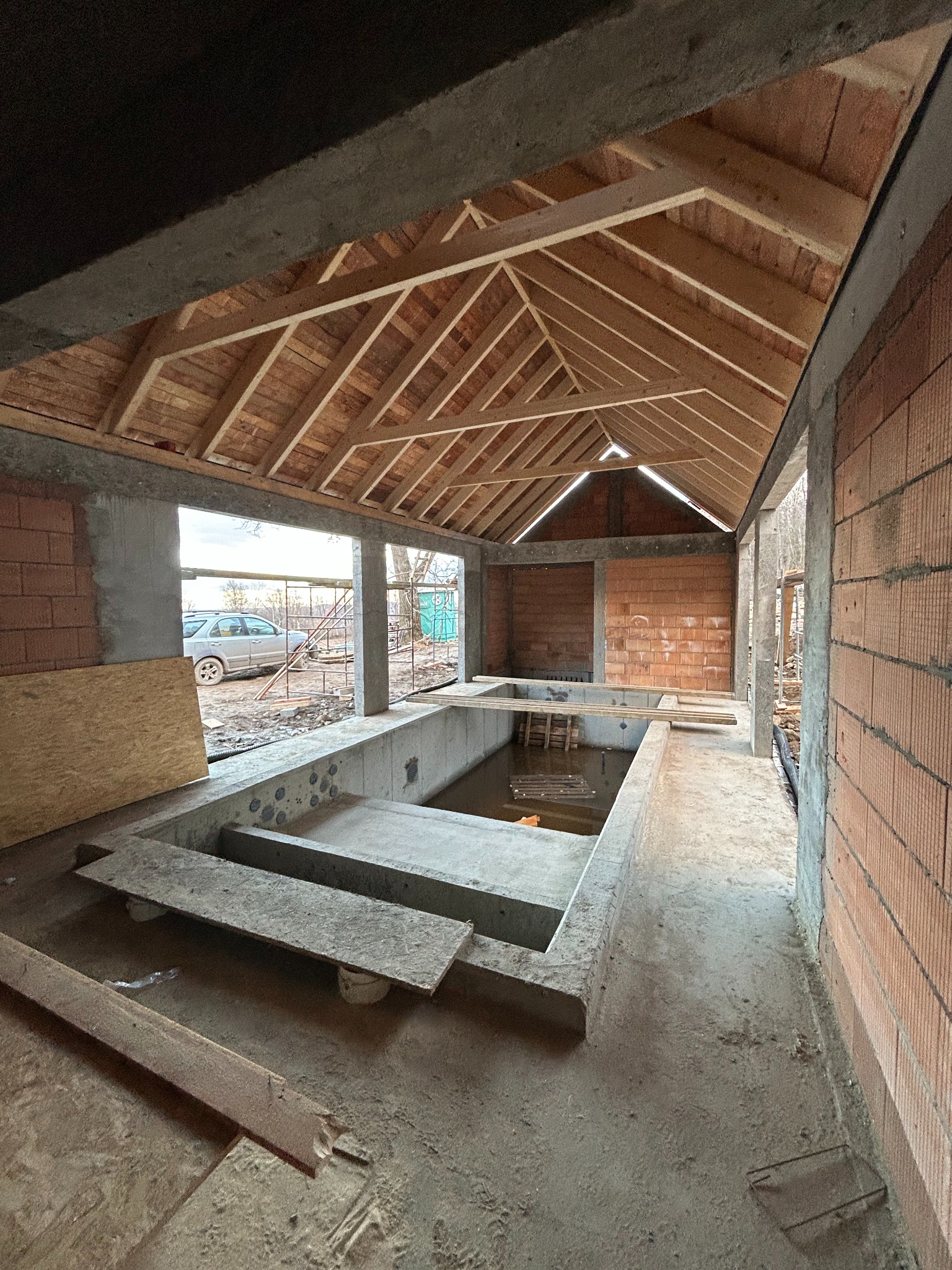Wnętrze budynku w trakcie budowy: surowe ściany z cegły, drewniana konstrukcja dachu, betonowy basen wypełniony wodą, widok na zewnątrz przez niezabudowane otwory okienne, samochód w tle.