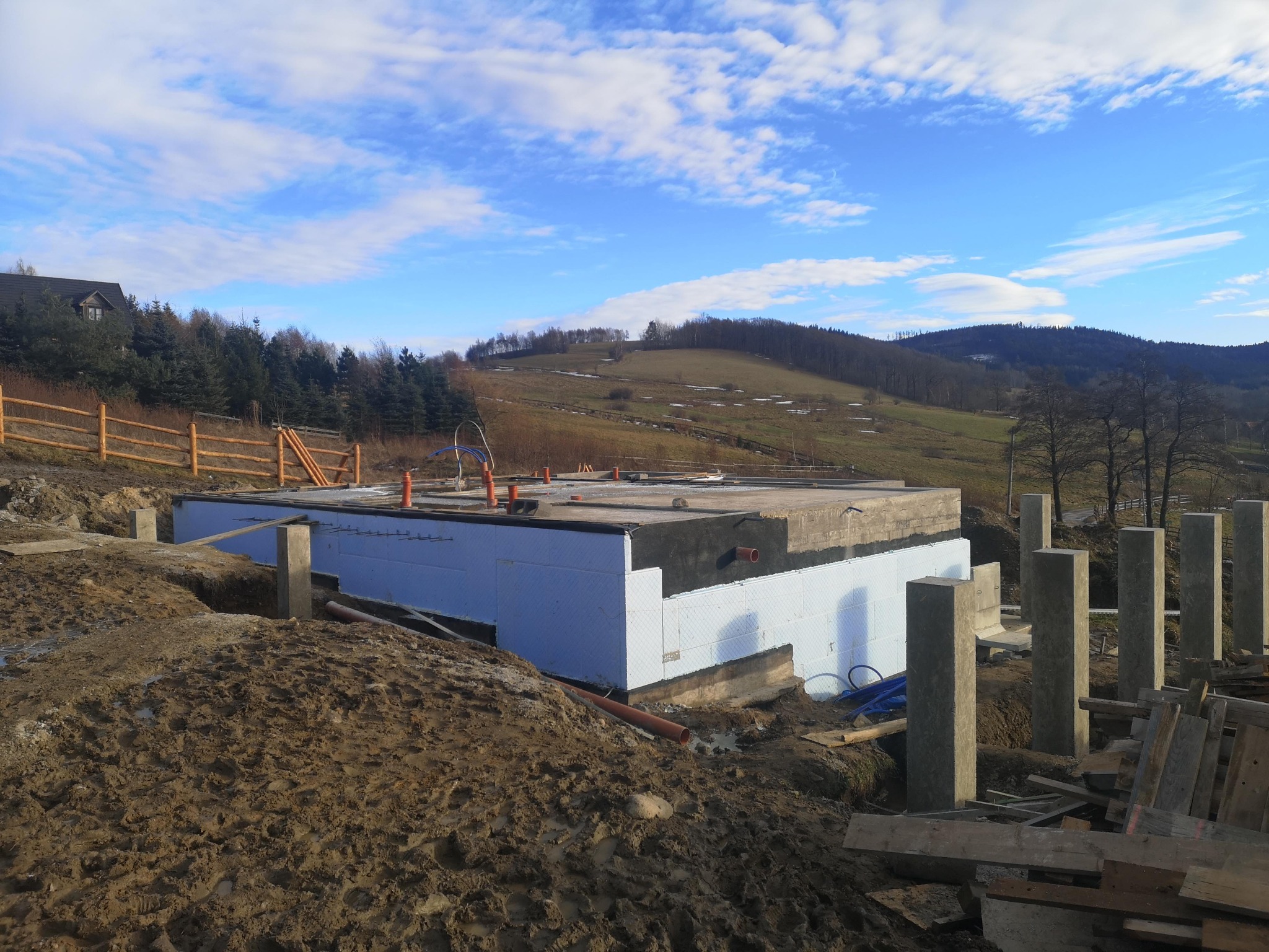 Fundament płyty budynku mieszkalnego, ocieplony styropianem, z widocznymi pionowymi słupami i elementami szalunków, na tle pagórkowatego krajobrazu z lasami i błękitnym niebem z chmurami.