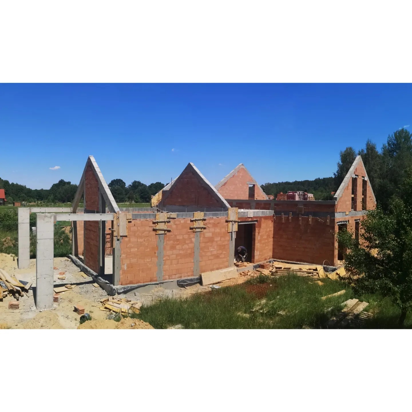 Wznoszenie ścian budynku mieszkalnego z czerwonej cegły ceramicznej, widoczne betonowe słupy nośne i zbrojenie, teren budowy z fragmentami zieleni i błękitnym niebem w tle.