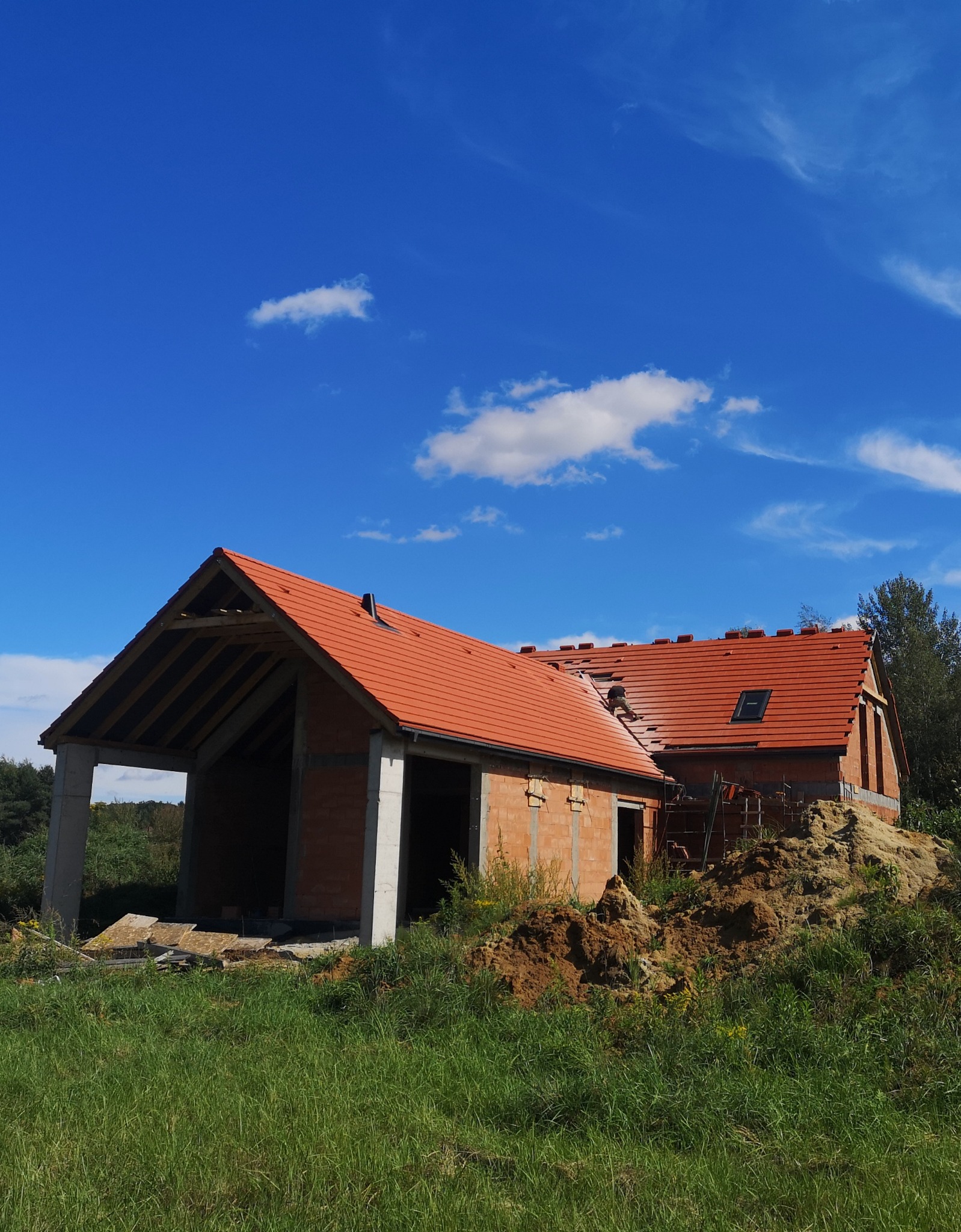 Stan surowy otwarty domu z czerwonym dachem, widoczne pustaki i betonowe słupy, na dachu pracownik układa dachówkę, w tle błękitne niebo z chmurami.