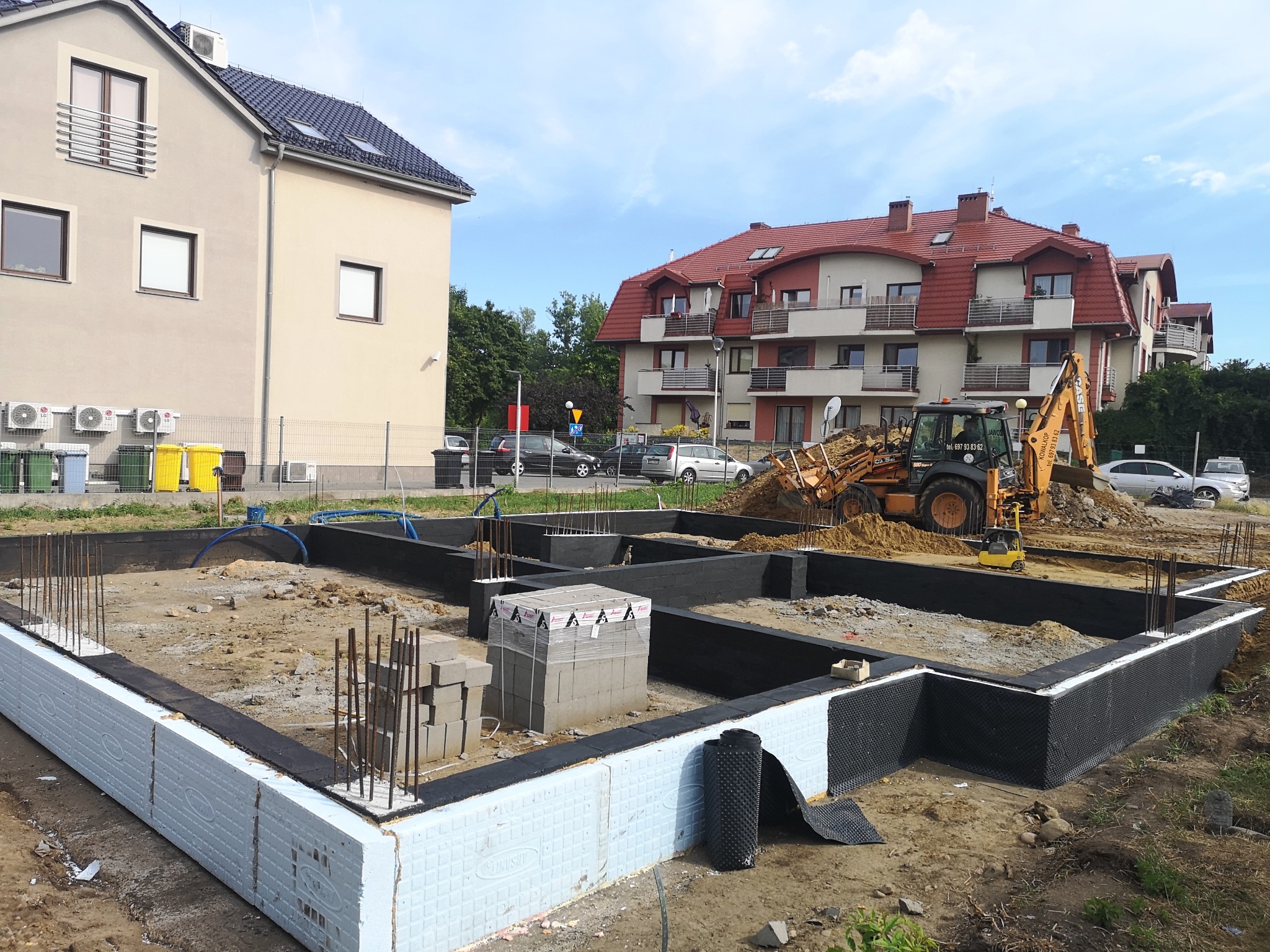 Budowa fundamentów domu w Pieszycach, widoczne izolowane ściany fundamentowe, wystające pręty zbrojeniowe i koparka w tle.