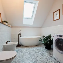 Jasna łazienka na poddaszu z białą wanną wolnostojącą, bidetem, toaletą, pralką Hotpoint Ariston i wzorzystą szaro-białą podłogą, oświetlona oknem dachowym.
