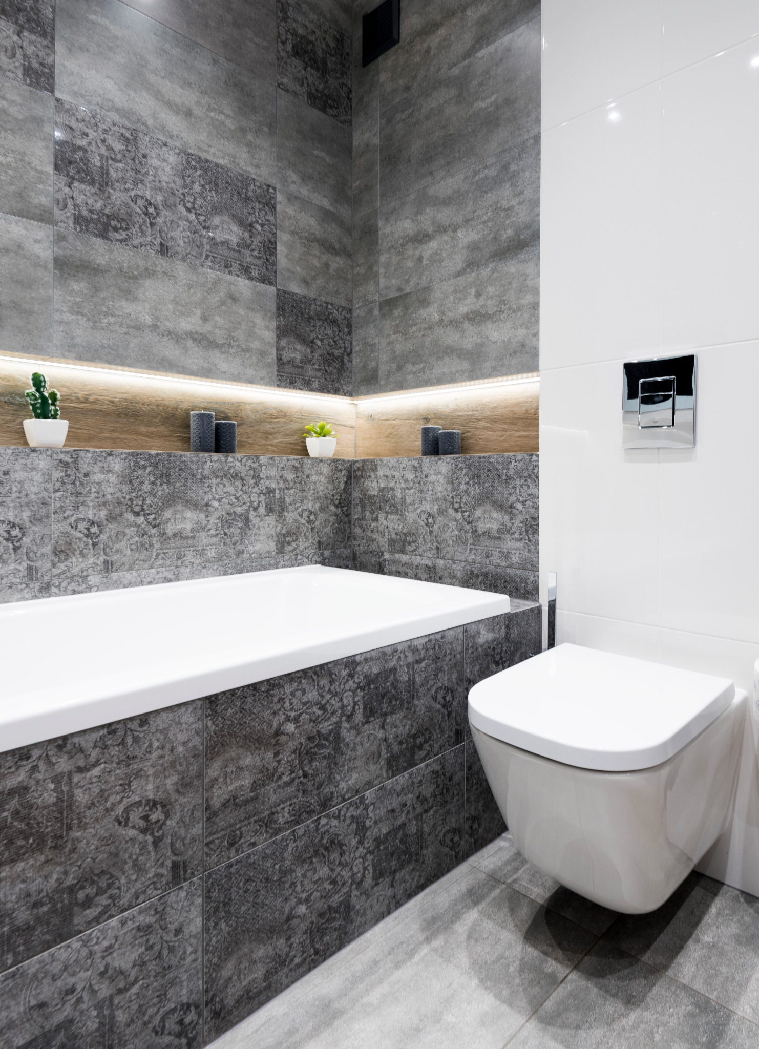 Nowoczesna łazienka z szarymi płytkami o wzorzystej fakturze, białą wanną i podwieszaną toaletą, oświetlona półka z dekoracjami roślinnymi i świecami, chromowany przycisk spłuczki.