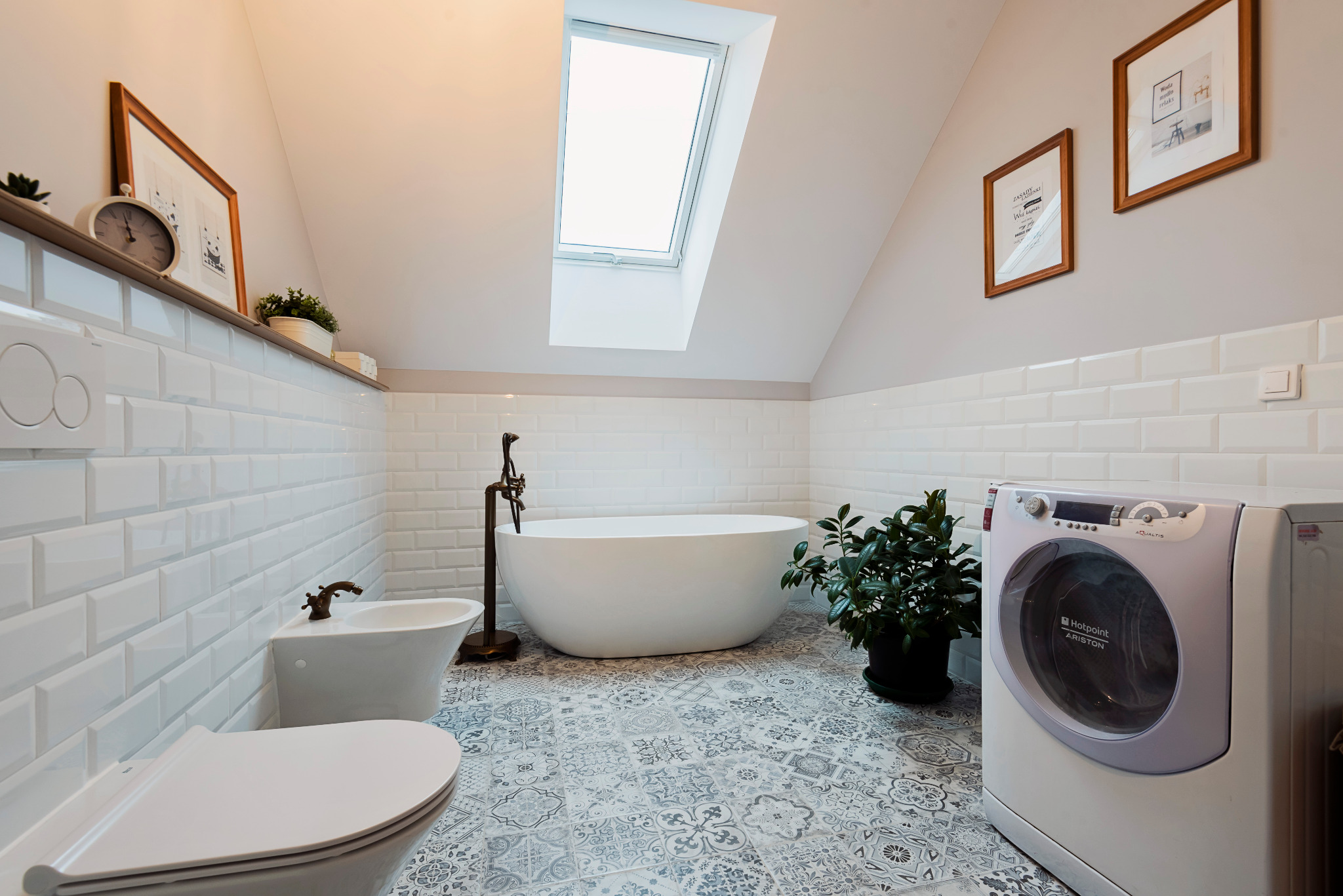 Jasna łazienka na poddaszu z białą wanną wolnostojącą, bidetem, toaletą, pralką Hotpoint Ariston i wzorzystą szaro-białą podłogą, oświetlona oknem dachowym.