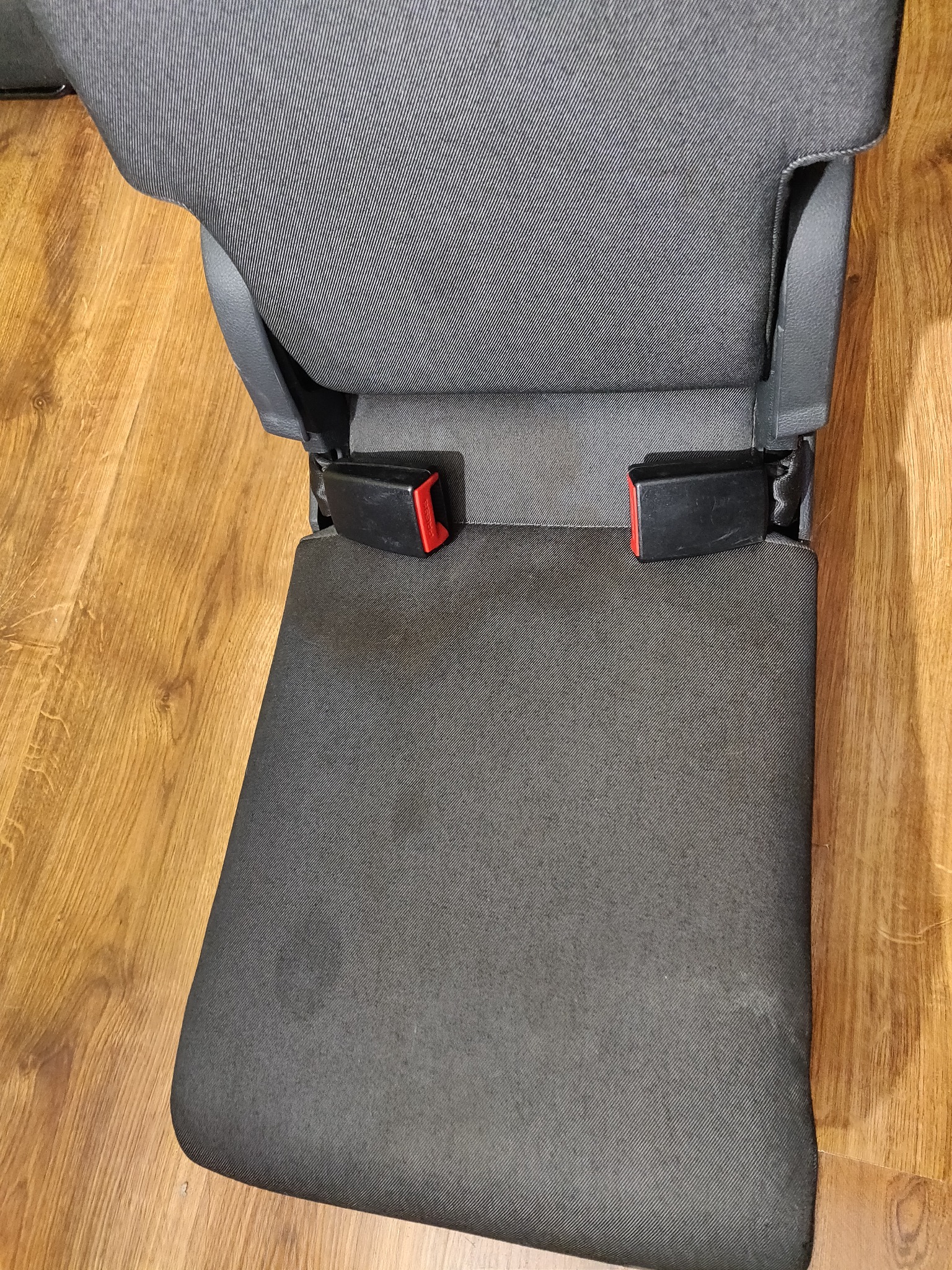 Szare, tapicerowane siedzenie samochodowe z widocznymi pasami bezpieczeństwa, ułożone na drewnianej podłodze.