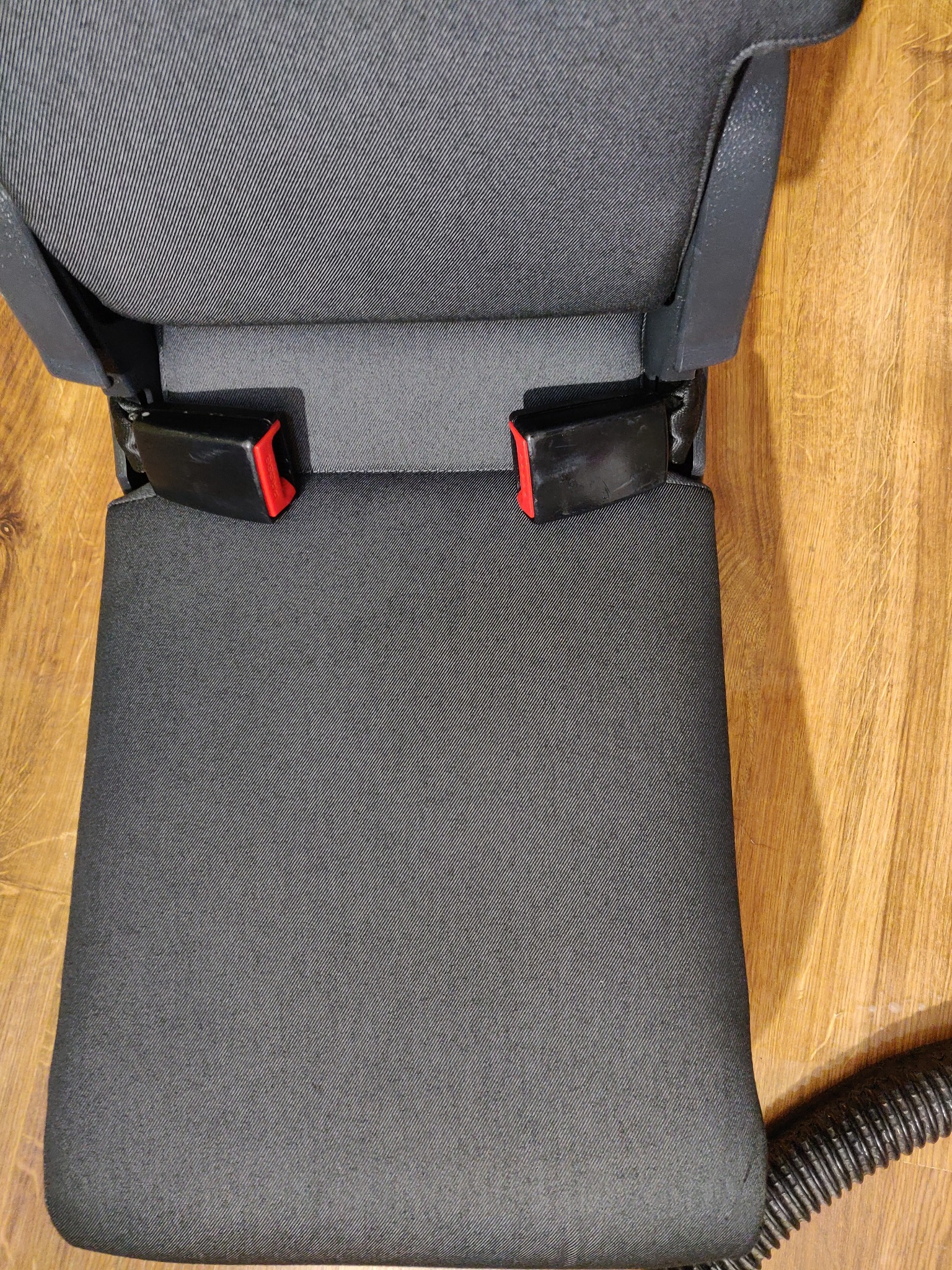 Czarny fotel samochodowy z widocznymi zapięciami pasów bezpieczeństwa na tle drewnianej podłogi, fragment węża odkurzacza w prawym dolnym rogu.