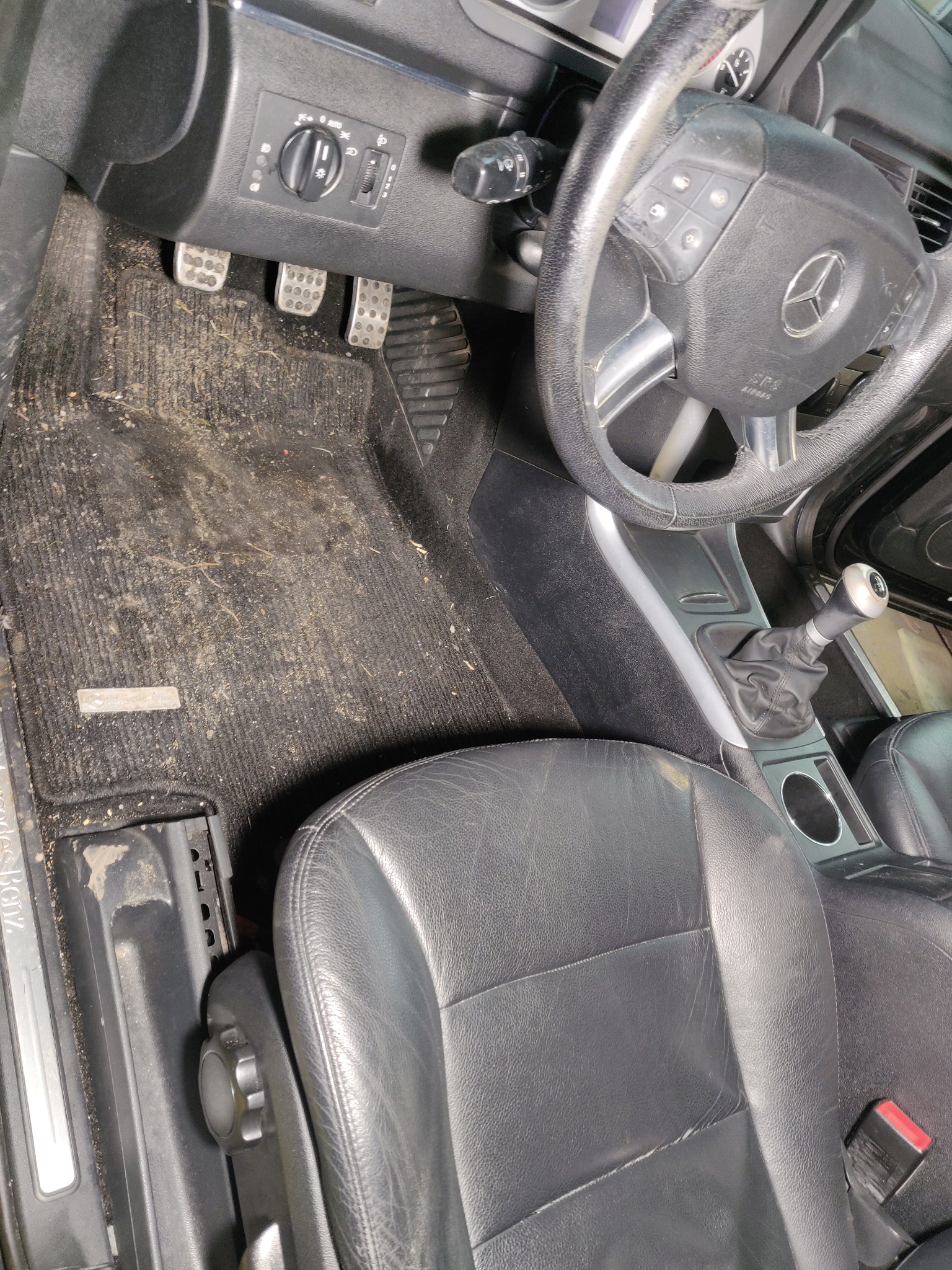 Wnętrze brudnego samochodu Mercedes-Benz przed czyszczeniem, widoczny zabrudzony dywanik podłogowy, skórzany fotel z przetarciami i kierownica.