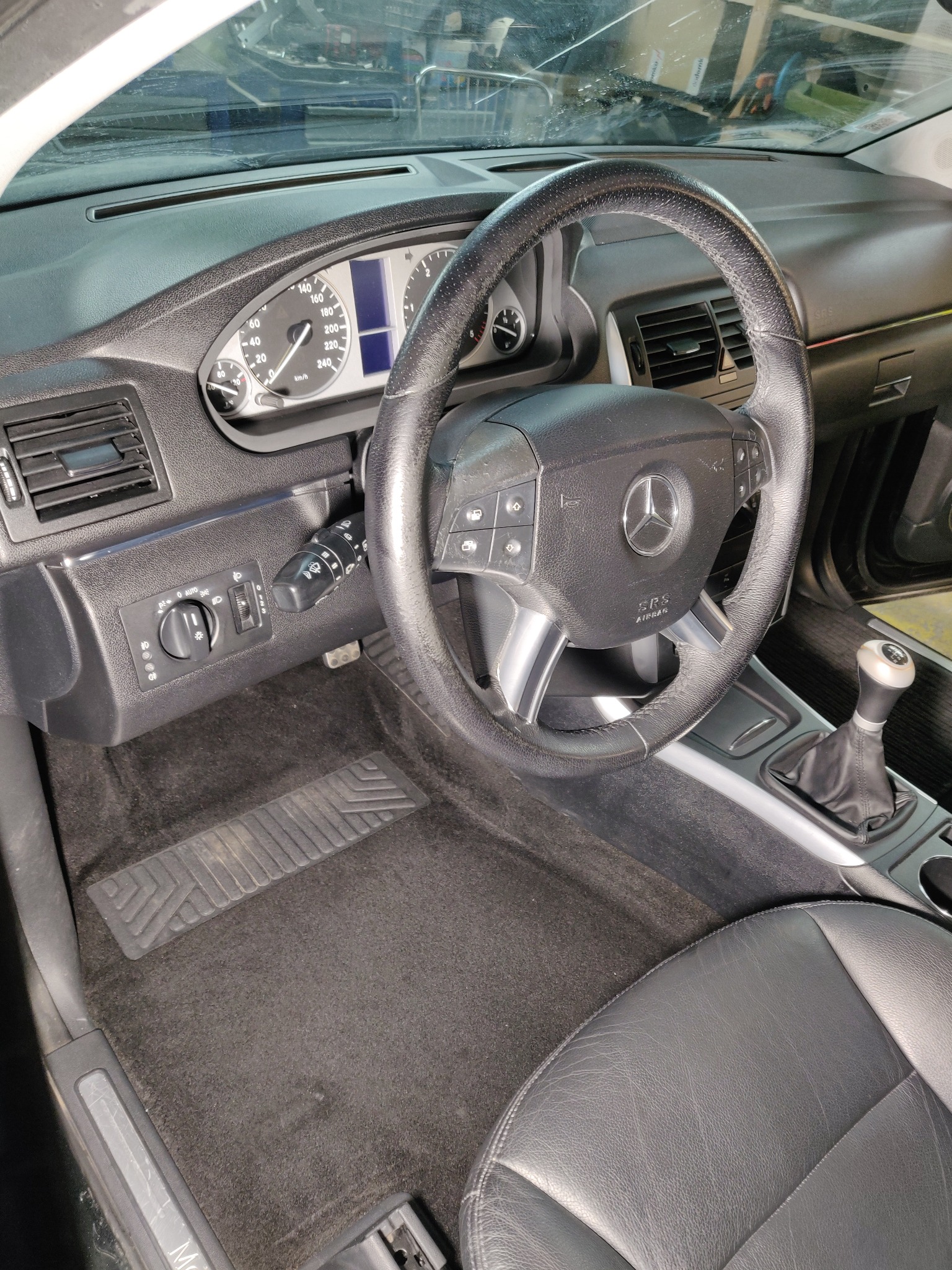Wnętrze samochodu marki Mercedes-Benz z widoczną deską rozdzielczą, kierownicą z logo, dźwignią zmiany biegów i czarną tapicerką. Dywaniki wymagają odświeżenia.