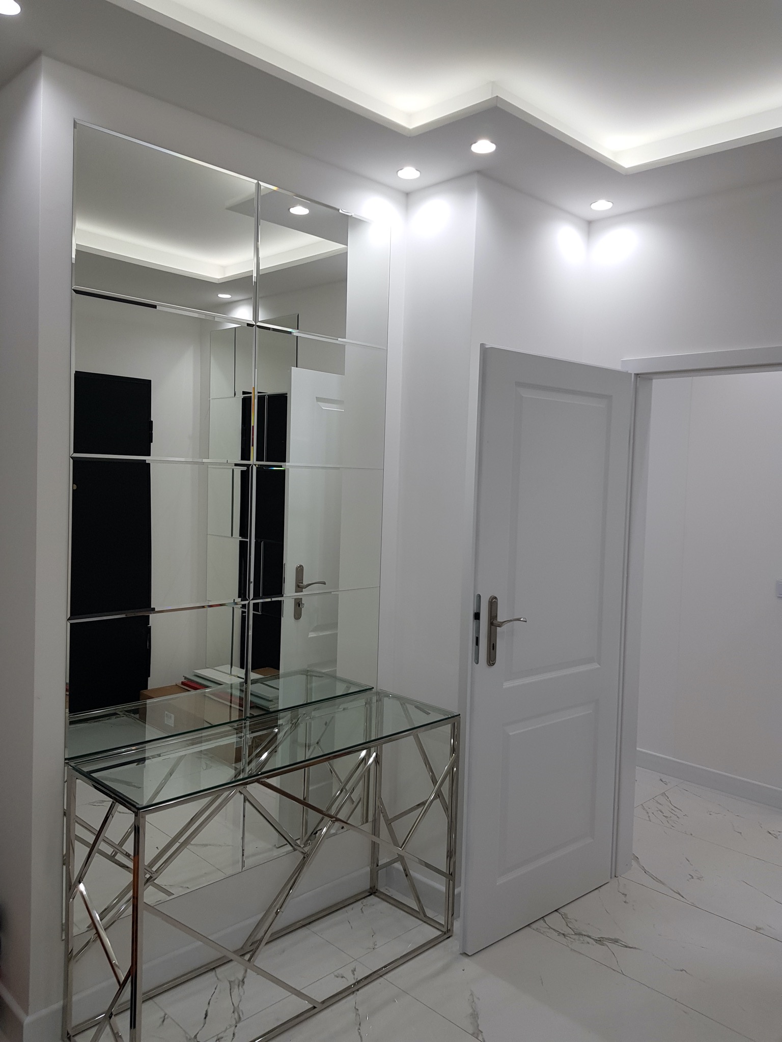 Nowoczesny przedpokój z lustrem złożonym z kwadratowych paneli, szklaną konsolą na chromowanej podstawie i białymi drzwiami.