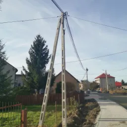 Betonowy słup telekomunikacyjny z przewodami na tle wiejskiej zabudowy i błękitnego nieba.