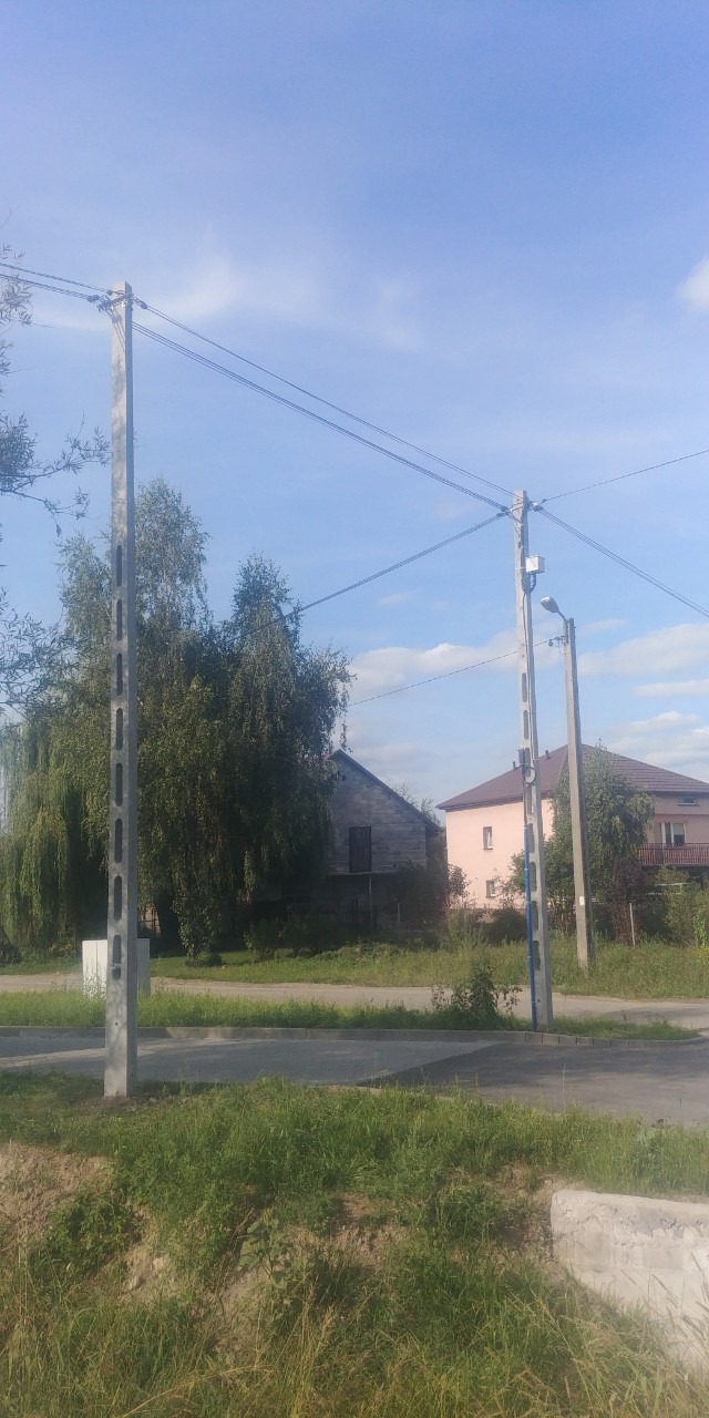 Betonowe słupy telekomunikacyjne z przewodami na tle nieba i zabudowań mieszkalnych.