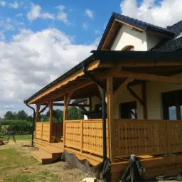 Drewniana weranda z ażurową balustradą przy domu z ciemną dachówką na tle błękitnego nieba z chmurami, widoczne narzędzia i materiały budowlane.