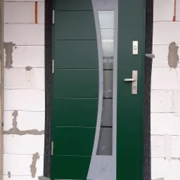 Nowoczesne, zielone drzwi wejściowe z przeszkleniem, zamontowane w surowym murze z betonu komórkowego, widoczne ślady zaprawy i pianki montażowej.