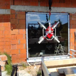 Montaż okna z użyciem przyssawki próżniowej na tle surowej ściany z cegły dziurawki, widoczne narzędzia i materiały budowlane.