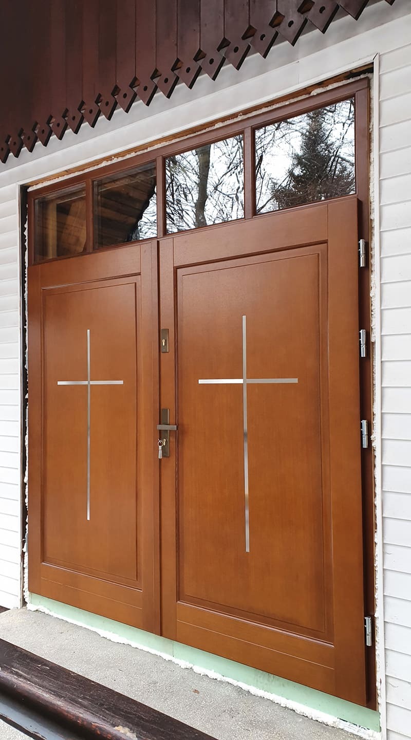 Brązowe, dwuskrzydłowe drzwi wejściowe z przeszkleniem górnym, ozdobione metalowymi krzyżami, zamontowane w białej elewacji z drewnianą podbitką.