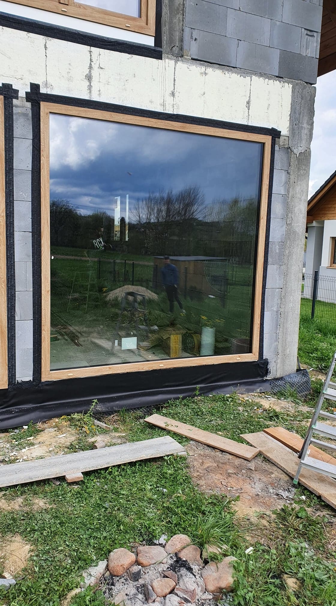 Montaż dużego okna z drewnianą ramą w surowym budynku z betonu komórkowego, widoczne uszczelnienie i odbicie osoby na zewnątrz w szybie.