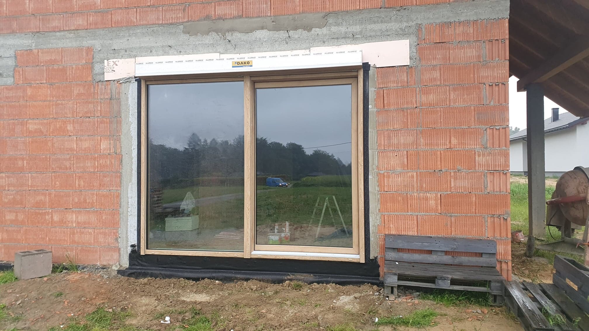 Montaż okna drewnianego DAKO w surowym murze z cegły, widoczne materiały izolacyjne i odbicie otoczenia w szybach.
