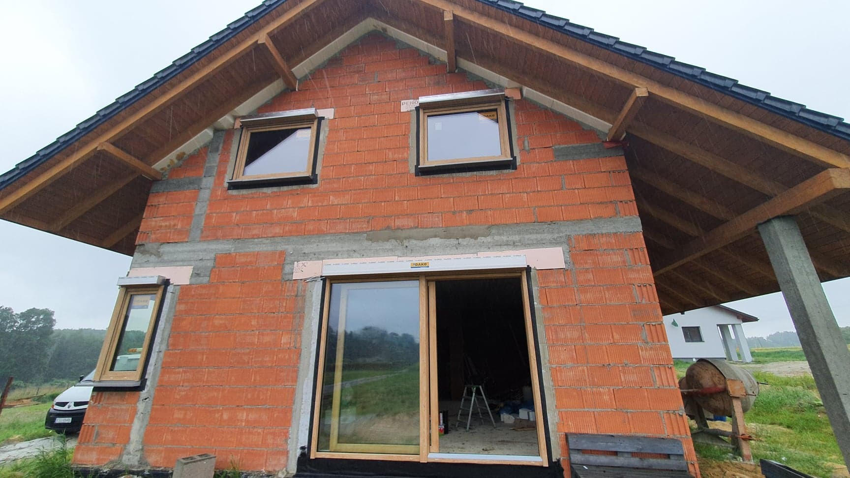 Surowy stan budynku w trakcie montażu stolarki okiennej i rolet zewnętrznych, widoczne okna z drewnopodobną ramą, częściowo zamontowane rolety z widocznym logo producenta na piance montażowej...