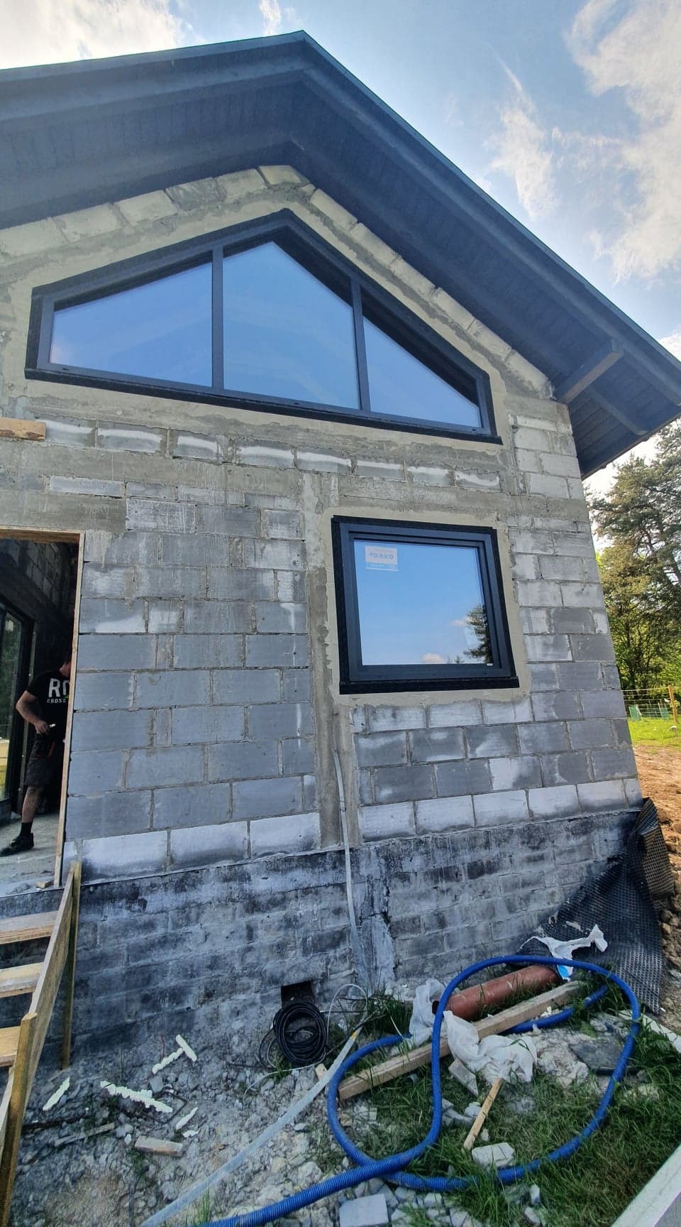 Montaż okien w surowym budynku z betonu komórkowego, widoczne okno trójkątne i prostokątne z czarnymi ramami, na dole nieporządek budowlany.