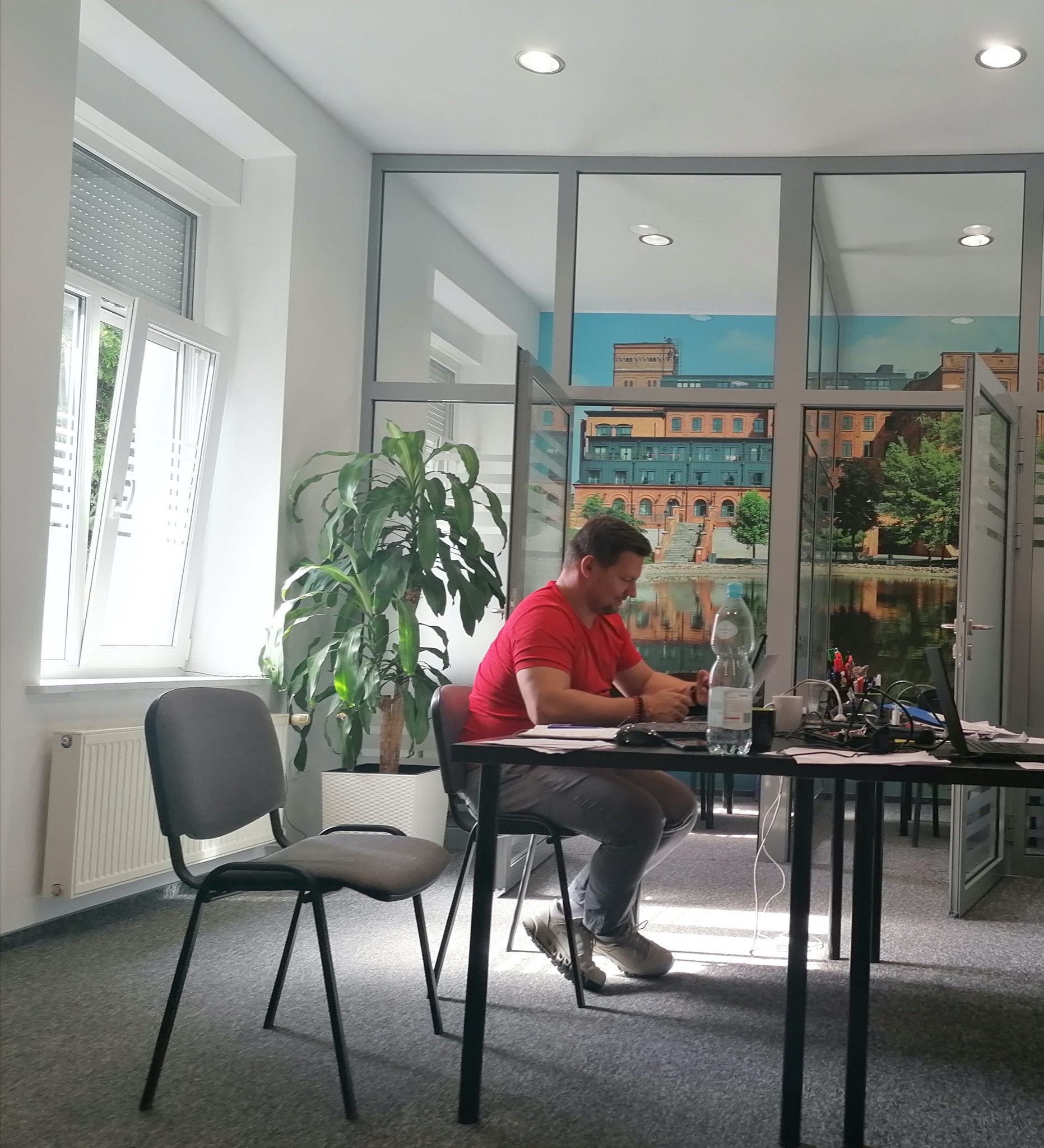 Mężczyzna w czerwonej koszulce siedzi przy biurku w nowoczesnym biurze, pracując na laptopie. W tle widoczne okno z widokiem na budynek przemysłowy i jezioro, a także roślina doniczkowa i puste...