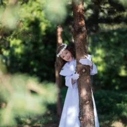 Dziewczynka w białej sukience i wianku z kwiatów, opierająca się o drzewo podczas sesji fotograficznej w plenerze.