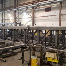 Wnętrze hali produkcyjnej z widoczną konstrukcją stalową w trakcie montażu, spawarka ESAB w tle, elementy transportowe na pierwszym planie.