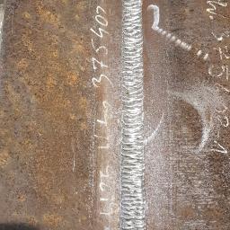 Jaśkiewicz Constructions - Spoina spawalnicza wykonana na zardzewiałej blasze stalowej, widoczne białe napisy techniczne i fragment butów spawacza.