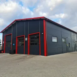 Nowoczesna hala z czarną elewacją z płyty warstwowej i czerwonymi akcentami wykończeniowymi, widoczne trzy bramy segmentowe i stos opon przed jedną z nich.