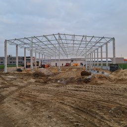 Jaśkiewicz Constructions - Szkielet konstrukcji stalowej hali w trakcie budowy, widoczne słupy i kratownica dachu, teren budowy z maszynami w tle, pochmurne niebo.