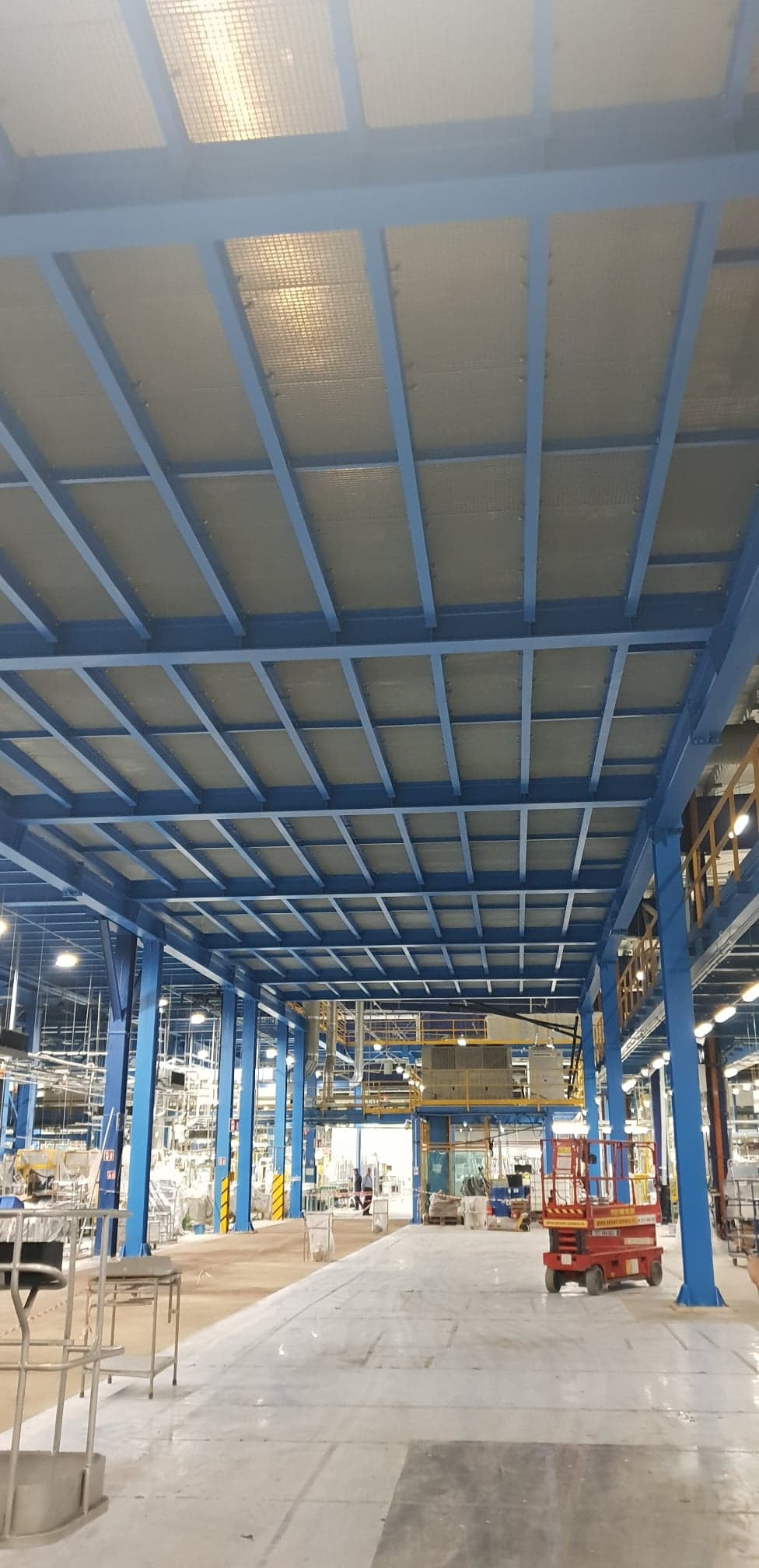 Wnętrze hali produkcyjnej w Śremie z widoczną konstrukcją stalową podtrzymującą sufit z paneli, z podnośnikiem nożycowym w tle i elementami instalacji pod stropem.