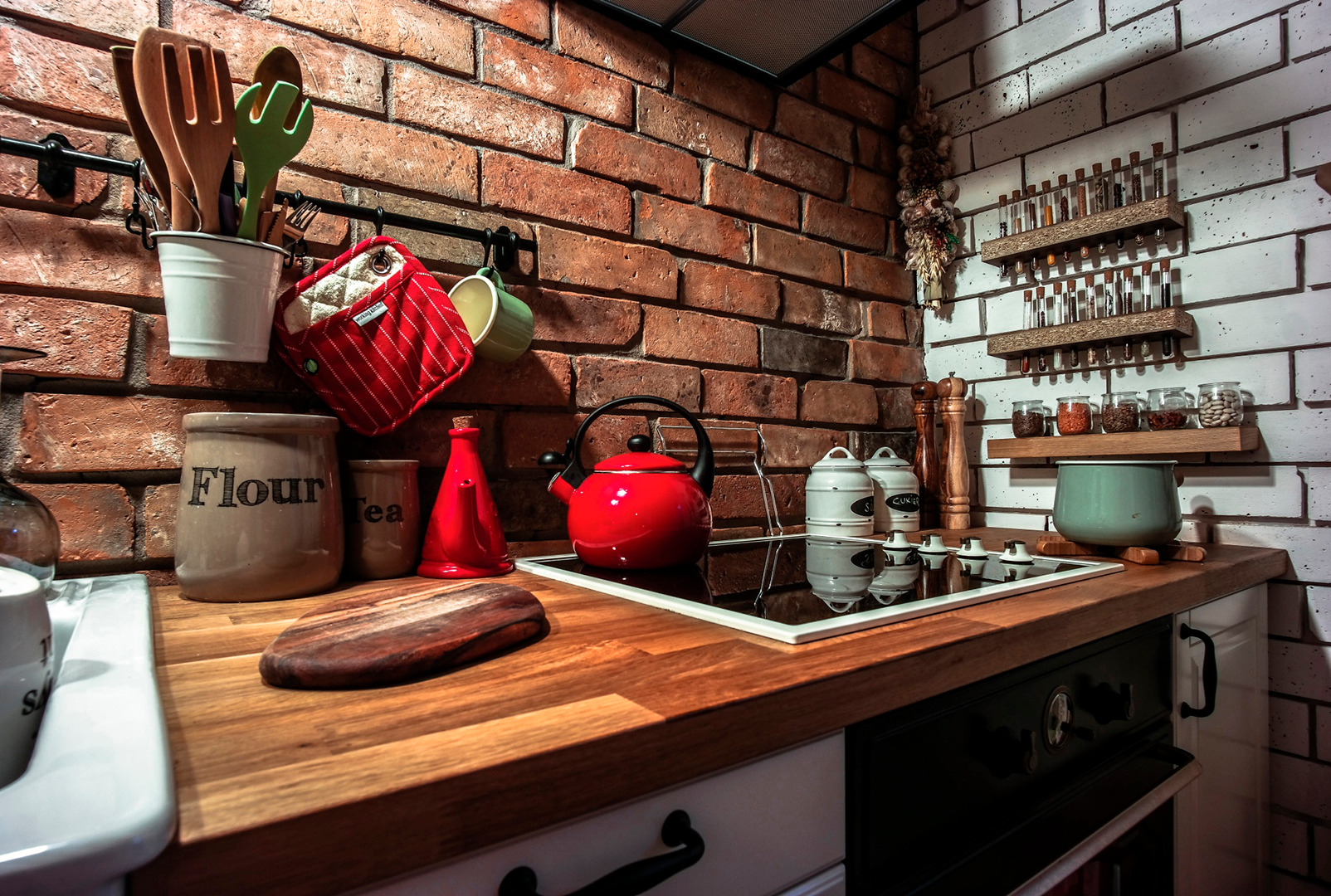 Rustykalna kuchnia z ceglaną ścianą, drewnianym blatem, czerwonym czajnikiem i przyprawami w probówkach na półkach.
