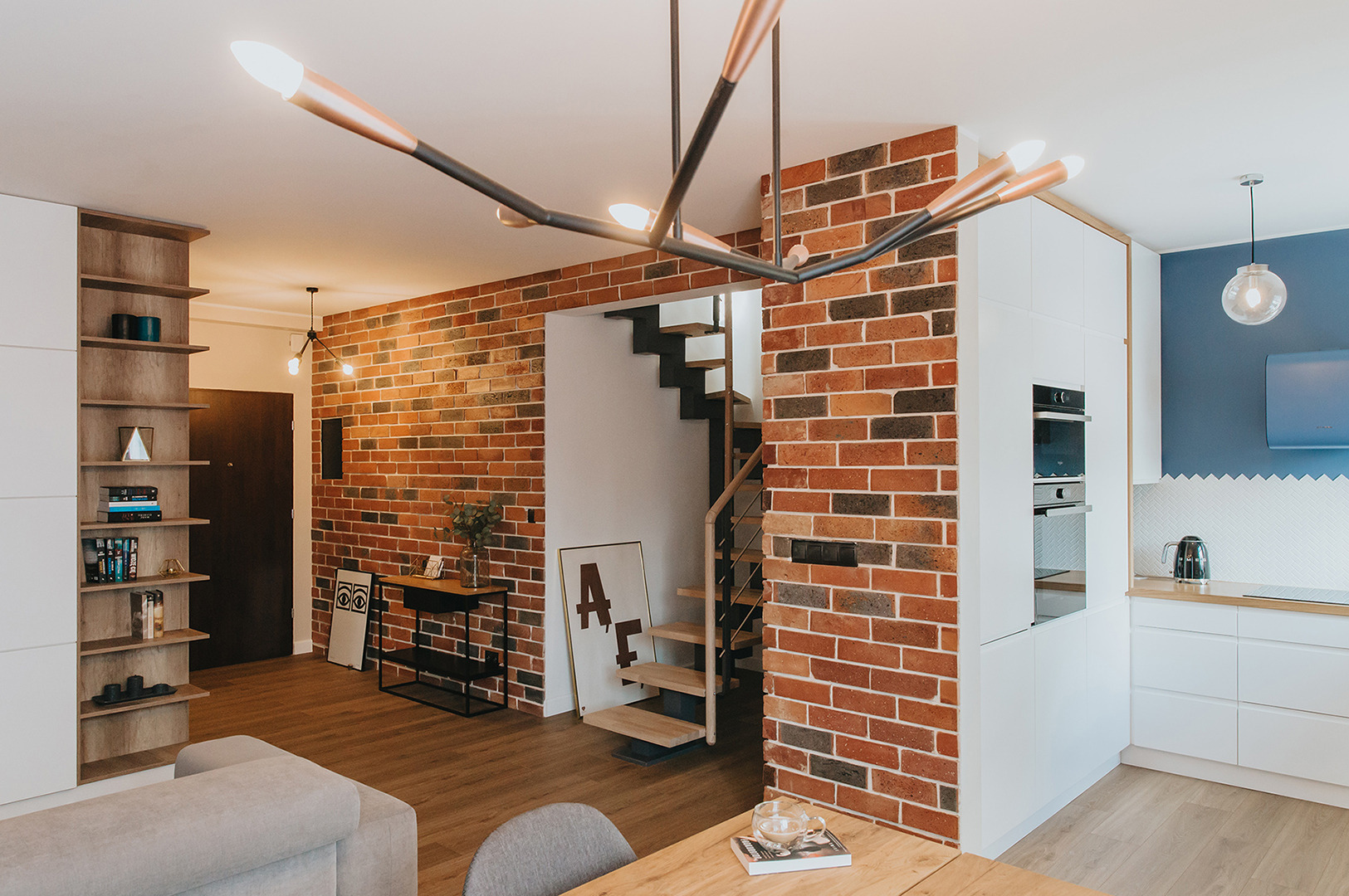 Nowoczesne wnętrze z ceglaną ścianą, drewnianymi schodami i designerską lampą sufitową, łączące elementy loftowe i skandynawskie.