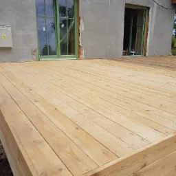 Nowo wybudowany taras z jasnego drewna, przylegający do szarego budynku z zielonymi oknami, widoczne niedokończone wykończenia elewacji i ziemia wokół.