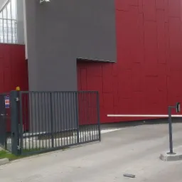 Szary metalowy szlaban automatyczny z lampą ostrzegawczą na tle czerwono-szarego budynku i metalowej furtki wejściowej.