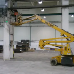 Pracownik w pomarańczowej kamizelce na podnośniku koszowym HA15IP, montujący element instalacji pod sufitem hali przemysłowej, widoczne rury, oświetlenie i konstrukcja wsporcza.