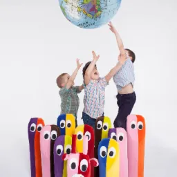 Trójka dzieci podskakuje, próbując dosięgnąć dmuchanego globu, otoczona przez rząd kolorowych, pluszowych maskotek z dużymi oczami.