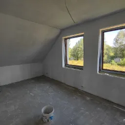 Poddasze po szpachlowaniu ścian i sufitu, widoczne dwa okna z widokiem na zieleń, na podłodze ślady po szpachli i wiadro po farbie.
