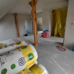 Wnętrze poddasza w trakcie prac wykończeniowych: widoczne bele konstrukcyjne, płyty karton-gips na ścianach, folia ochronna, oraz nieużyte rolki wełny mineralnej w białych opakowaniach z żółtymi...