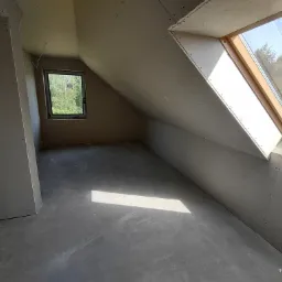 Poddasze w trakcie wykańczania, ściany i skośny sufit obłożone płytami kartonowo-gipsowymi, widoczne wkręty, okno dachowe z drewnianą ramą, betonowa podłoga z refleksem światła.