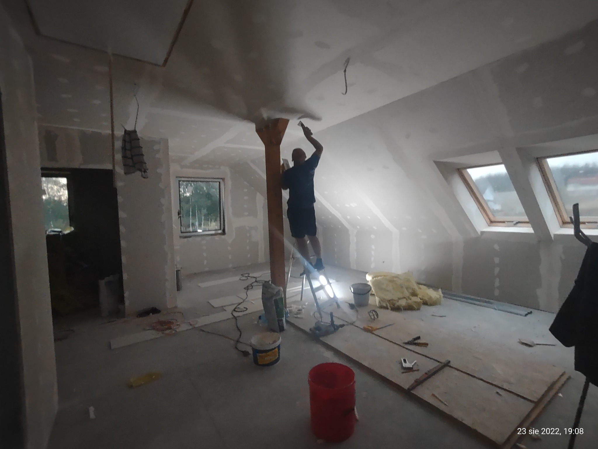 Pracownik na drabinie szpachluje sufit poddasza z oknami dachowymi, widoczne płyty kartonowo-gipsowe, narzędzia i materiały budowlane w nieuporządkowanym wnętrzu.