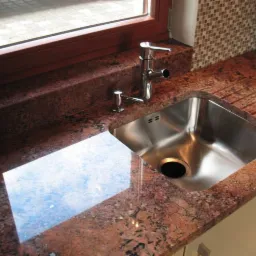 Błyszczący, stalowy zlew kuchenny wbudowany w czerwony, nakrapiany blat granitowy, z widocznym oknem i mozaikową ścianą w tle.