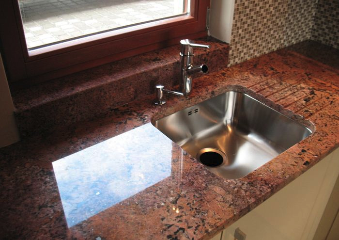Błyszczący, stalowy zlew kuchenny wbudowany w czerwony, nakrapiany blat granitowy, z widocznym oknem i mozaikową ścianą w tle.