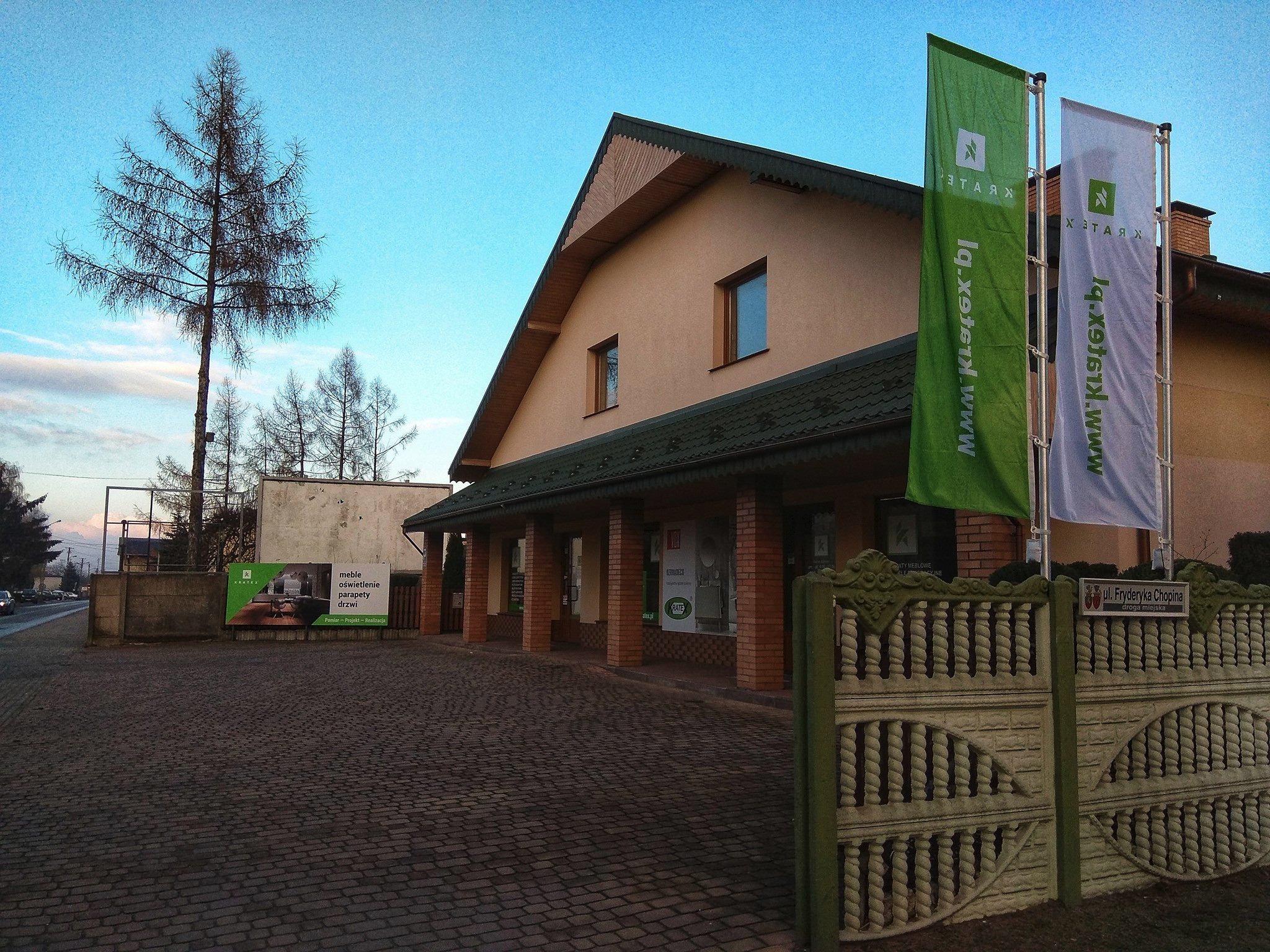 Fasada budynku firmy z branży wyposażenia wnętrz, z widocznymi flagami reklamowymi, ogrodzeniem z betonowych elementów oraz tablicą informacyjną z ofertą mebli, oświetlenia, parapetów i stolarki...