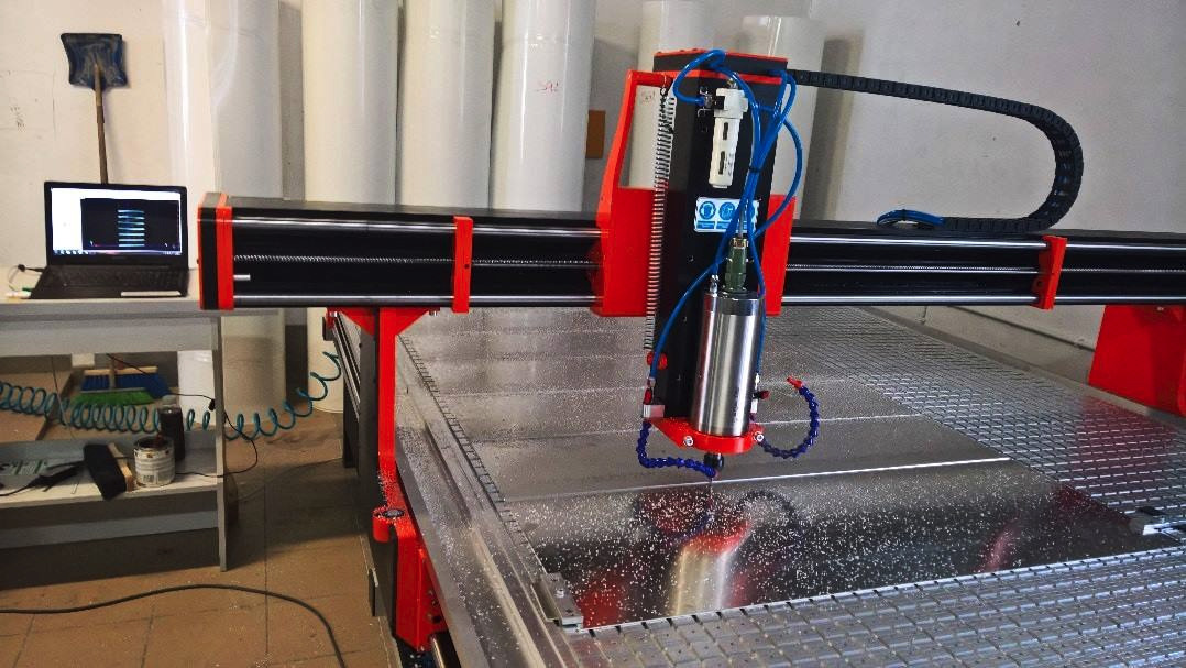 Frezarka CNC w Zduńskiej Woli w trakcie pracy, widoczne chłodziwo i opiłki na obrabianym materiale, obok laptop z oprogramowaniem sterującym.