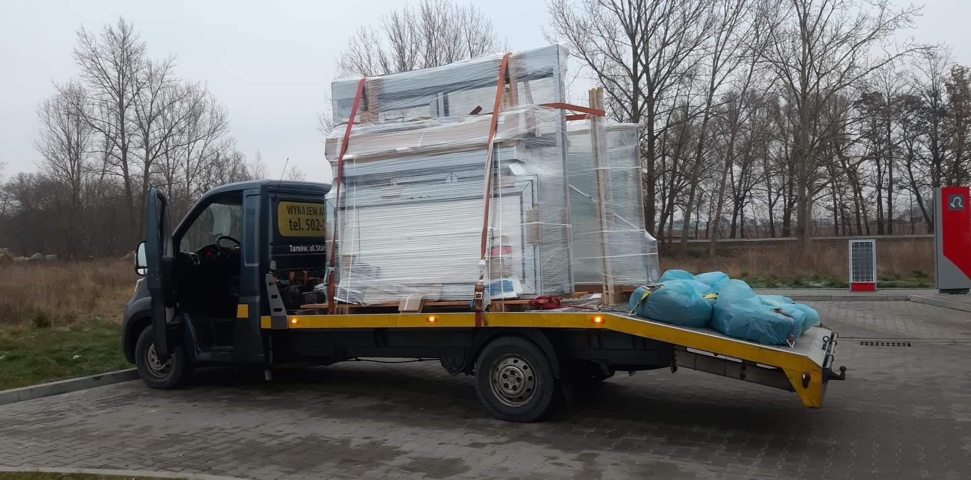 Laweta z otwartymi drzwiami kierowcy przewozi okna i drzwi owinięte folią, zabezpieczone pasami, na tle szarej, jesiennej scenerii.