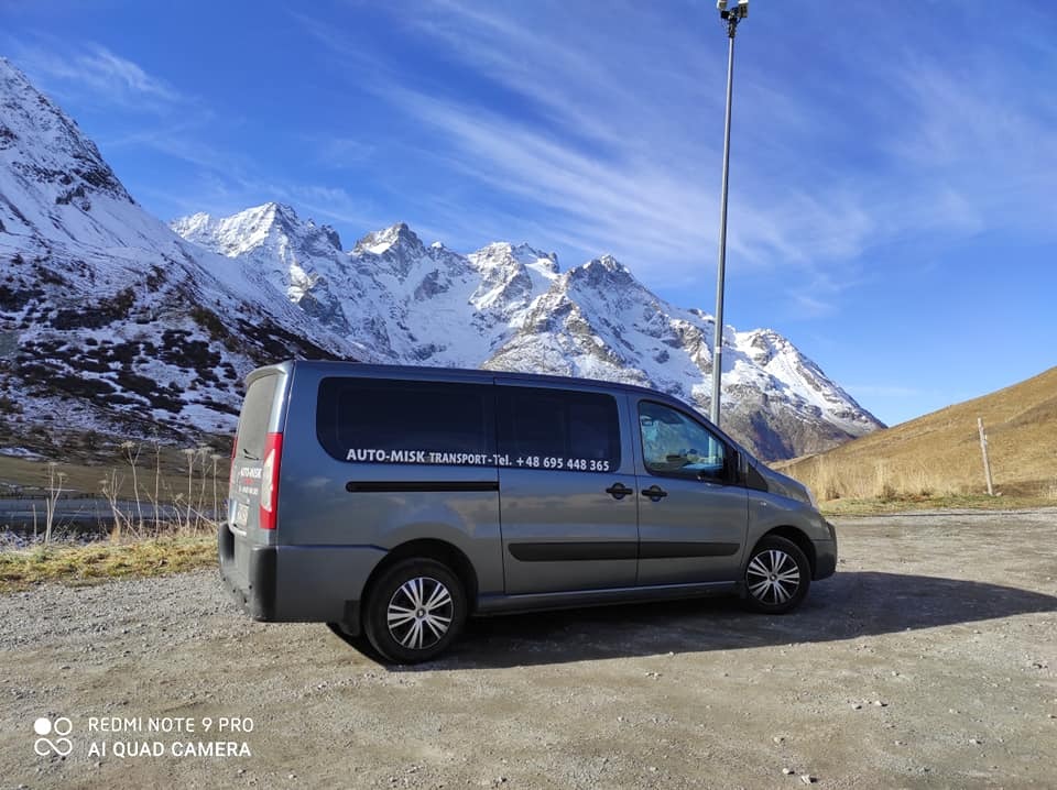 Szary bus transportowy z napisem AUTO-MISK TRANSPORT na tle górskiego krajobrazu ze śnieżnymi szczytami i błękitnym niebem.
