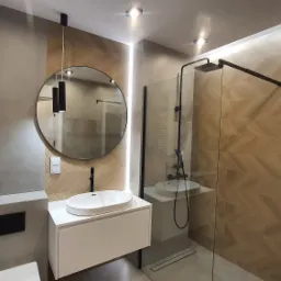 Nowoczesna łazienka z umywalką nablatową, okrągłym lustrem z podświetleniem LED, kabiną prysznicową typu walk-in i dekoracyjną ścianą z wzorem jodełki.