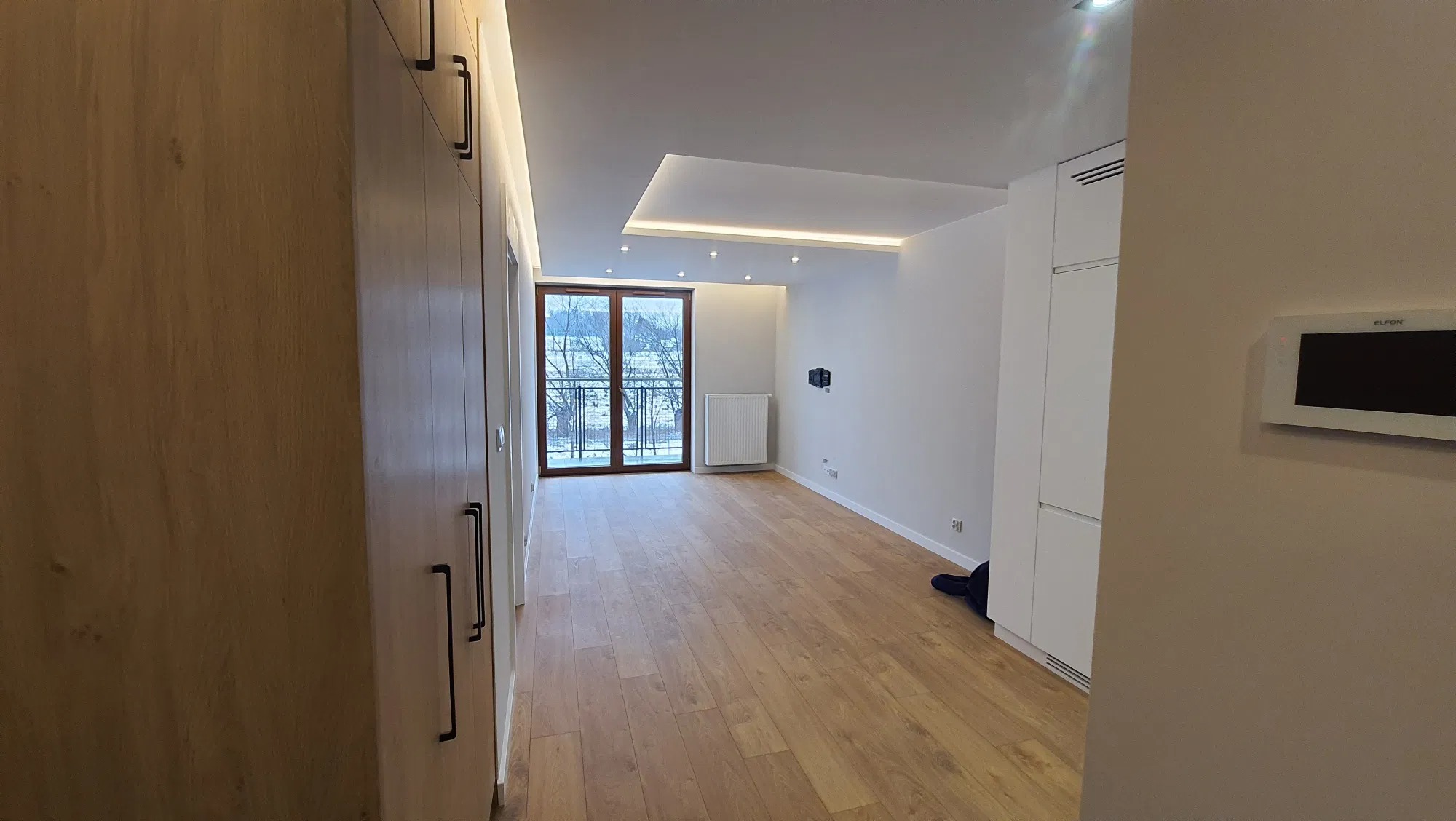 Jasne, nowoczesne wnętrze z drewnianą podłogą, białymi szafami w zabudowie i oknem balkonowym z widokiem na drzewa.